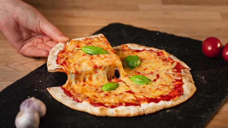 A hand pulling a slice of pizza topped with melted vegan mozzarella, with a stretchy cheese pull. The pizza is garnished with fresh basil leaves, and bright cherry tomatoes are seen in the background.