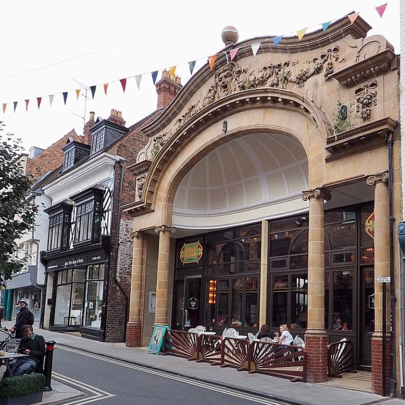 The image shows a historic building with an arched entrance and intricate stone carvings, featuring a café or restaurant called "Cosy Club." The facade is adorned with classical architectural details, while the street outside has decorative bunting, giving a festive feel to the scene. A few people are seated outside, enjoying the ambiance of the quaint, narrow street lined with other charming, old-style buildings.