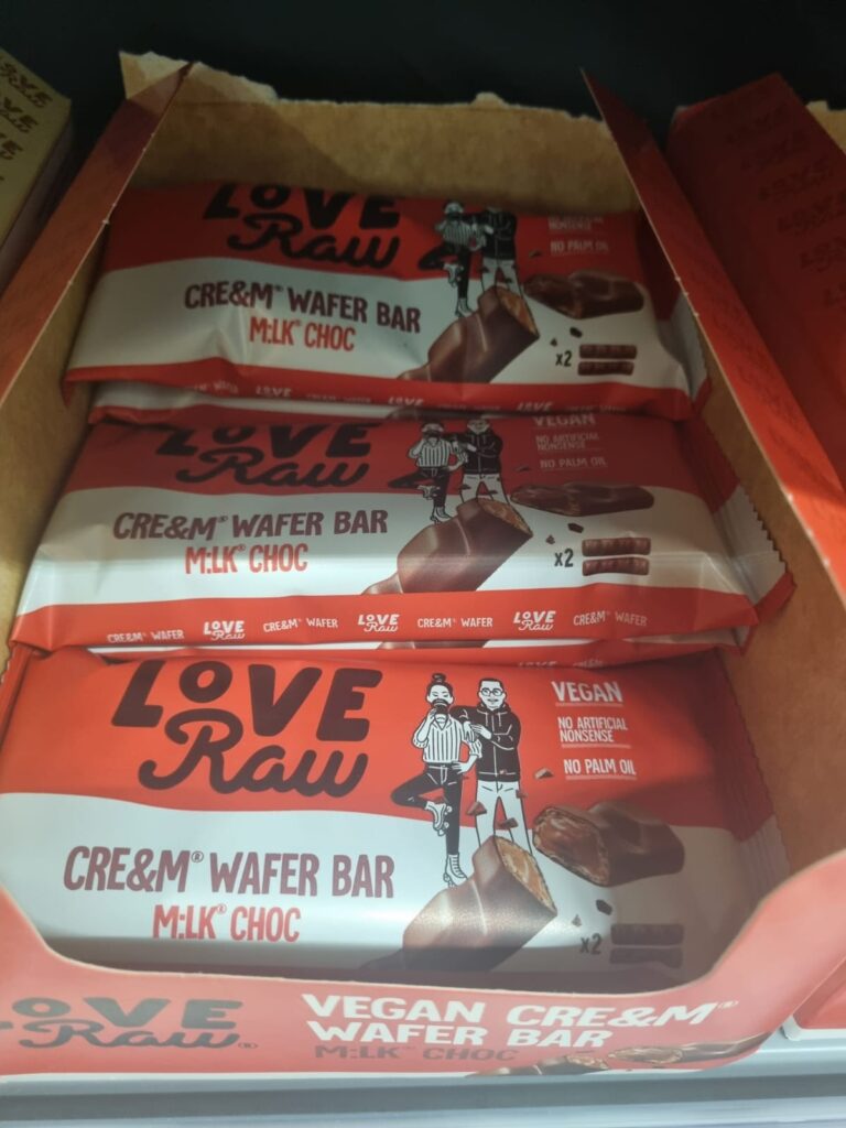 This image features a display of Love Raw's Vegan Cre&m Wafer Bars in "Milk Choc" flavour. The packaging highlights that the product is vegan, contains no artificial ingredients, and is free from palm oil. The wrapper includes an illustration of two characters and an image of the chocolate bar showing its creamy, wafer-filled layers. The package also states that there are two bars inside each pack.