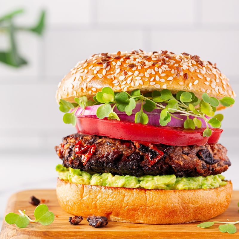A close-up of a plant-based burger with a sesame seed bun, layers of avocado spread, a veggie patty, tomato slices, red onion, and fresh microgreens.