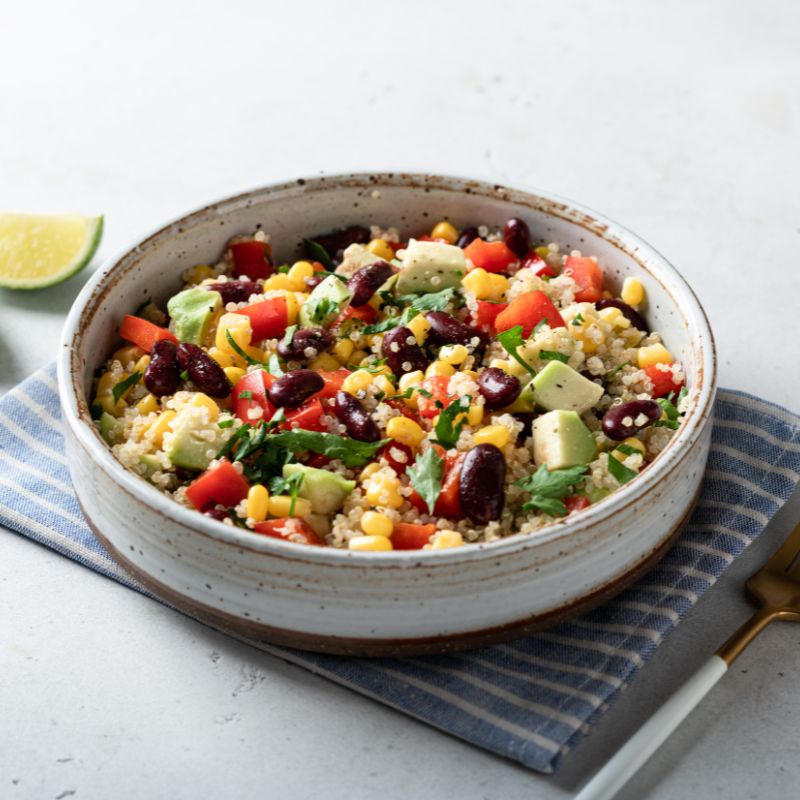 A healthy quinoa bowl featuring black beans, avocado, corn, diced red peppers, and cilantro, served in a rustic ceramic bowl. This colourful and protein-packed dish offers a convenient and nutritious meal option.