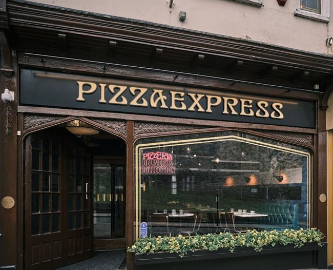  A view of the Pizza Express storefront with a dark wooden frame, ornate carvings, and a large sign displaying "PIZZAEXPRESS" above the entrance. The interior is dimly visible through the large window with a neon sign that reads "Pizzeria Delivery & Collection.