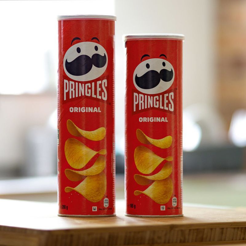 Two upright Pringles Original cans with red packaging, featuring the brand's iconic logo and an illustration of crisps, placed on a wooden surface in a bright room.