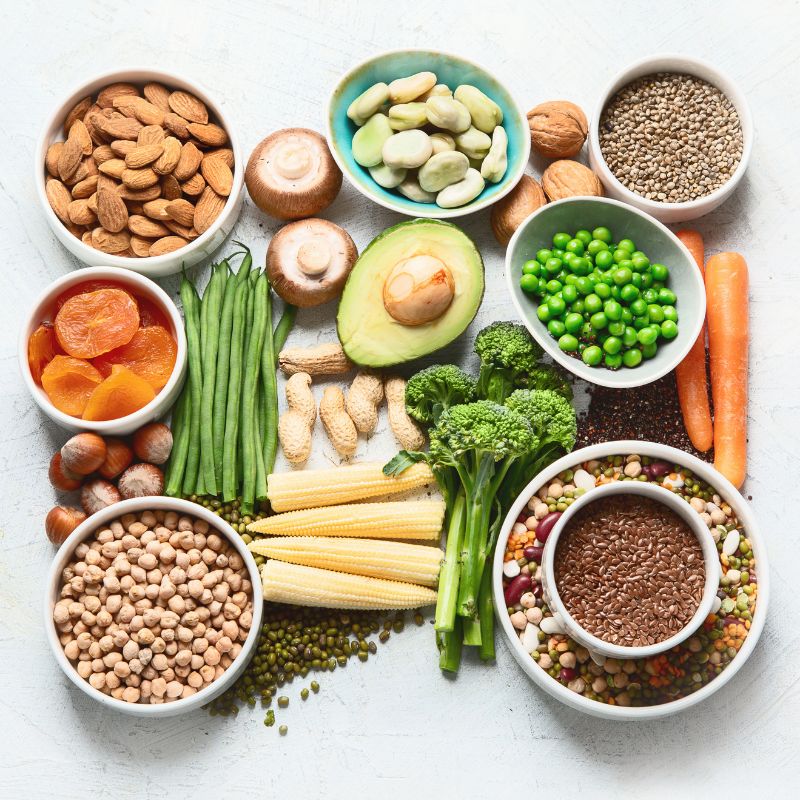 A variety of fresh vegetables, legumes, nuts, and seeds, including carrots, broccoli, avocado, green beans, chickpeas, and dried apricots, laid out in small bowls and piles.