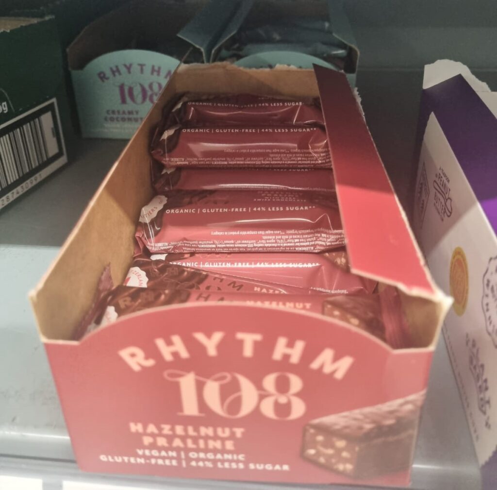 This image displays a box of Rhythm 108 Hazelnut Praline bars. The packaging highlights that the product is vegan, organic, gluten-free, and contains 44% less sugar. The bars are shown in a red wrapper with text emphasising their clean ingredients and reduced sugar content, alongside an image of the praline chocolate.