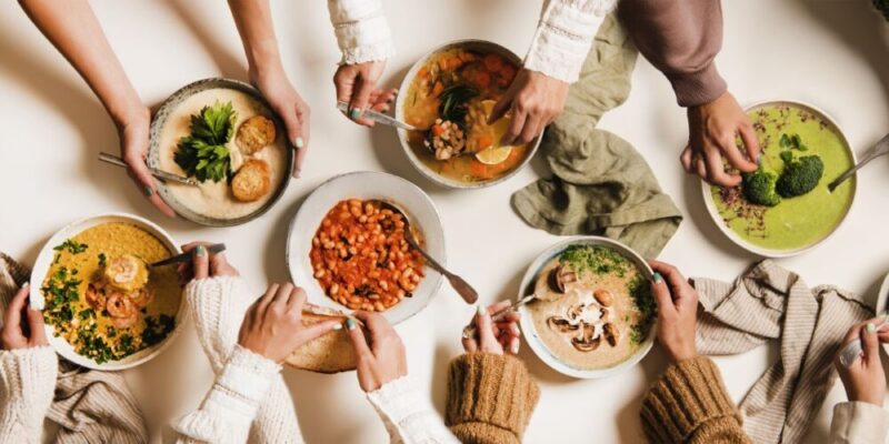 A top-down view of multiple hands reaching for bowls of various vegan soups and stews on a table. The dishes include vibrant green broccoli soup, hearty bean stew, creamy mushroom soup, and more, showcasing a cosy and communal meal setup.