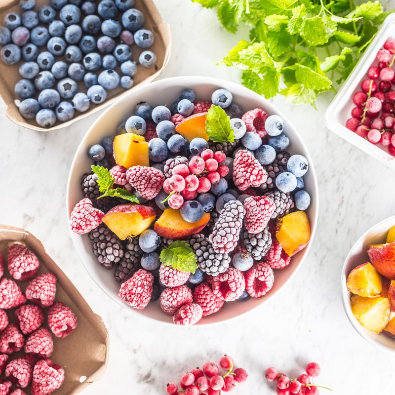 A collection of frozen mixed berries and peaches in bowls surrounded by fresh blueberries, raspberries, and mint leaves on a marble countertop.