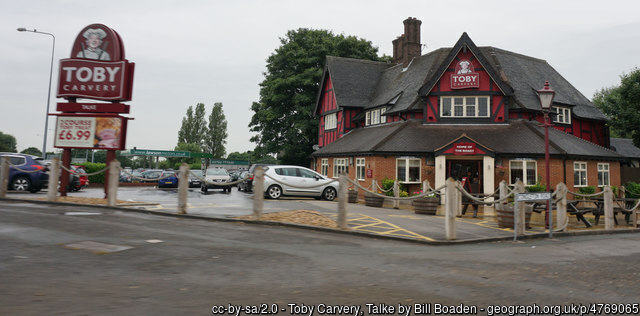 The image shows a Toby Carvery restaurant with a classic brick and timber exterior, situated in a parking area with a few cars parked nearby. A prominent sign outside advertises a meal price of £6.99, emphasising its affordability. The surroundings include trees and a slightly overcast sky, adding a casual atmosphere to the scene.