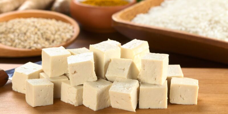 A pile of cubed tofu placed on a wooden surface. The tofu is light beige in colour with a smooth texture, and the cubes are neatly stacked. In the background, there are blurred bowls of grains and rice, adding context to the tofu as a key ingredient in a meal. The overall presentation highlights the tofu’s versatility in cooking.