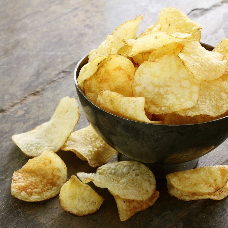 A rustic metal bowl filled with golden crips, with several crips scattered around the bowl on a weathered wooden surface. The image emphasises the texture and light golden-brown edges of the chips, suggesting a fresh, crunchy snack.