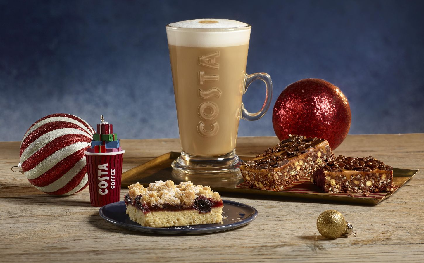 This image showcases a festive holiday setup from Costa Coffee. A frothy latte in a tall Costa glass is surrounded by an assortment of desserts, including a crumb-topped berry slice and chocolate-covered nut bars, all arranged alongside decorative holiday ornaments like a red and white striped bauble and a miniature Costa coffee cup ornament with stacked gifts. The scene exudes a cozy, festive atmosphere, highlighting seasonal treats.