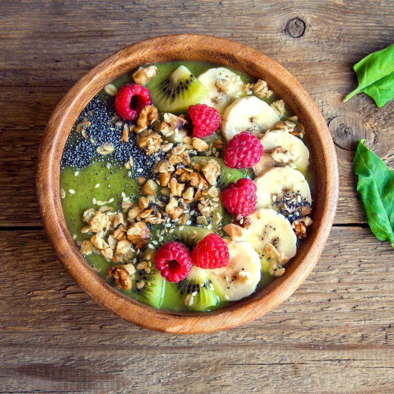 A wooden bowl filled with a vibrant green smoothie base with vegan protein, topped with banana slices, kiwi, raspberries, chia seeds, granola, and walnuts. The bowl is arranged beautifully on a wooden surface, offering a nutrient-dense and visually appealing meal.