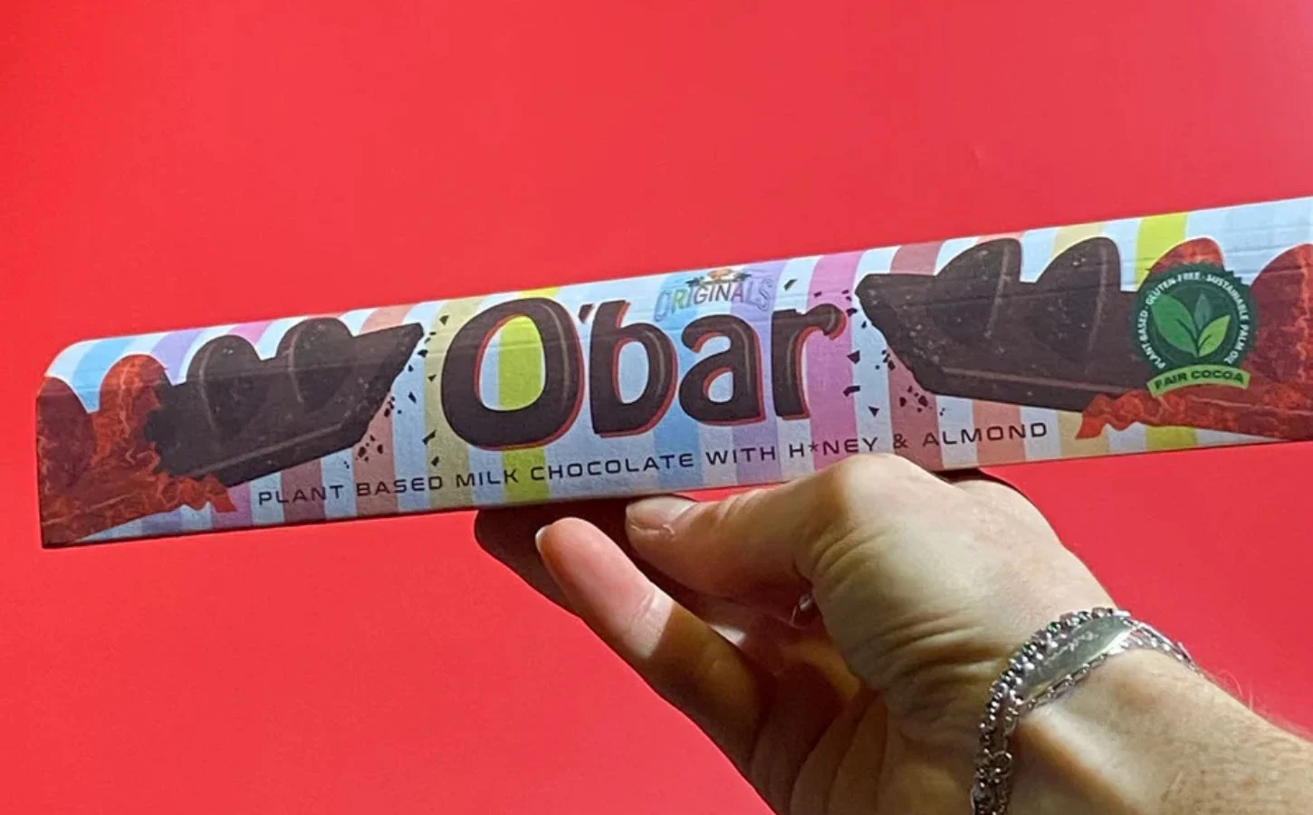 Image of Vegan Chocolate Bar Packaging: A hand holding a colorful, striped package labeled "Obar," which describes the contents as plant-based milk chocolate with honey and almond. The packaging includes a fair cocoa logo, indicating sustainable sourcing, with a vibrant red background.