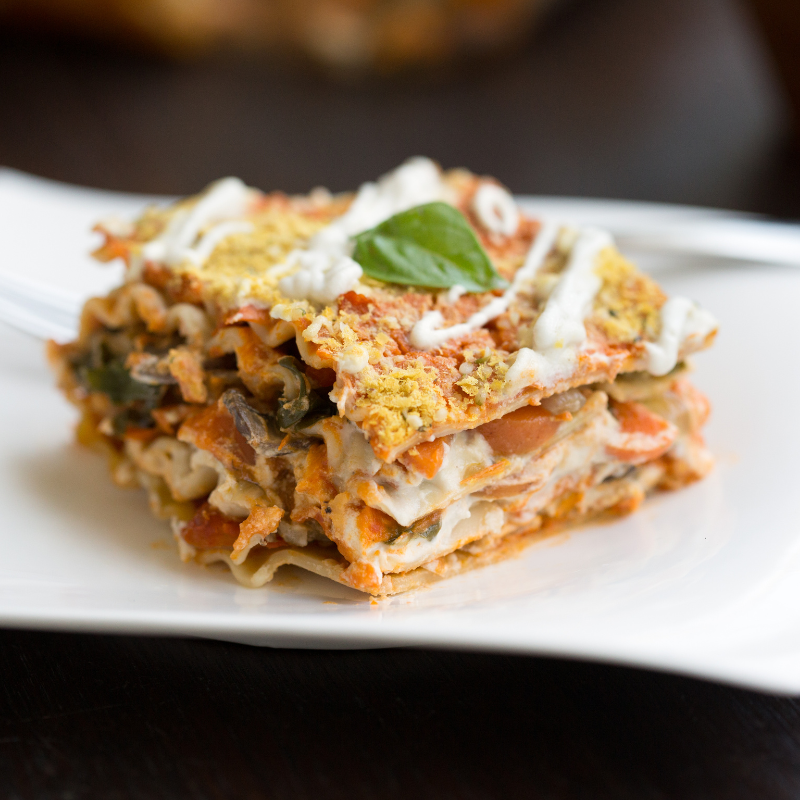 A slice of vegan lasagne made with tofu ricotta. The lasagne has layers of pasta, a rich tomato sauce, vegetables, and a creamy tofu-based ricotta filling. The top is garnished with a sprinkle of nutritional yeast, giving it a cheesy appearance, and a drizzle of white sauce, likely a cashew or vegan cream sauce. A fresh basil leaf is placed on top, adding a touch of green to the golden-brown dish. The lasagne looks hearty and flavourful, ideal for a comforting plant-based meal.