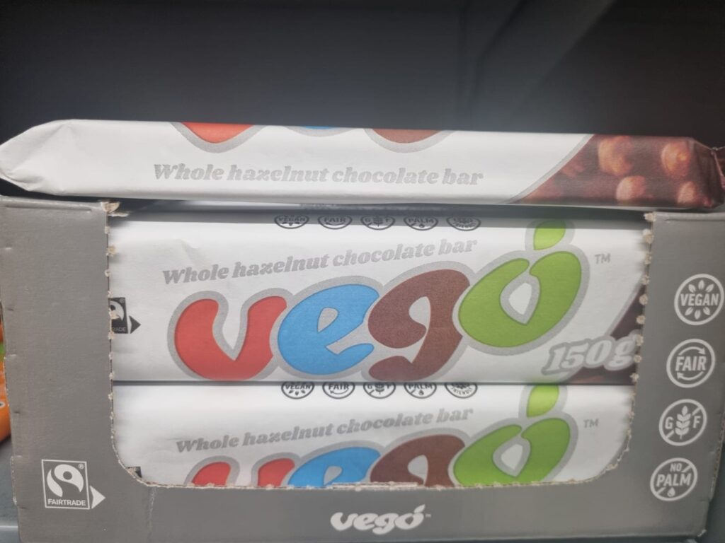 This image shows a display box containing Vego Whole Hazelnut Chocolate Bars, which are 150g each. The packaging highlights that the product is vegan, fair-trade certified, gluten-free, and palm oil-free. The Vego brand name is prominently featured in large, colourful lettering on the bars.
