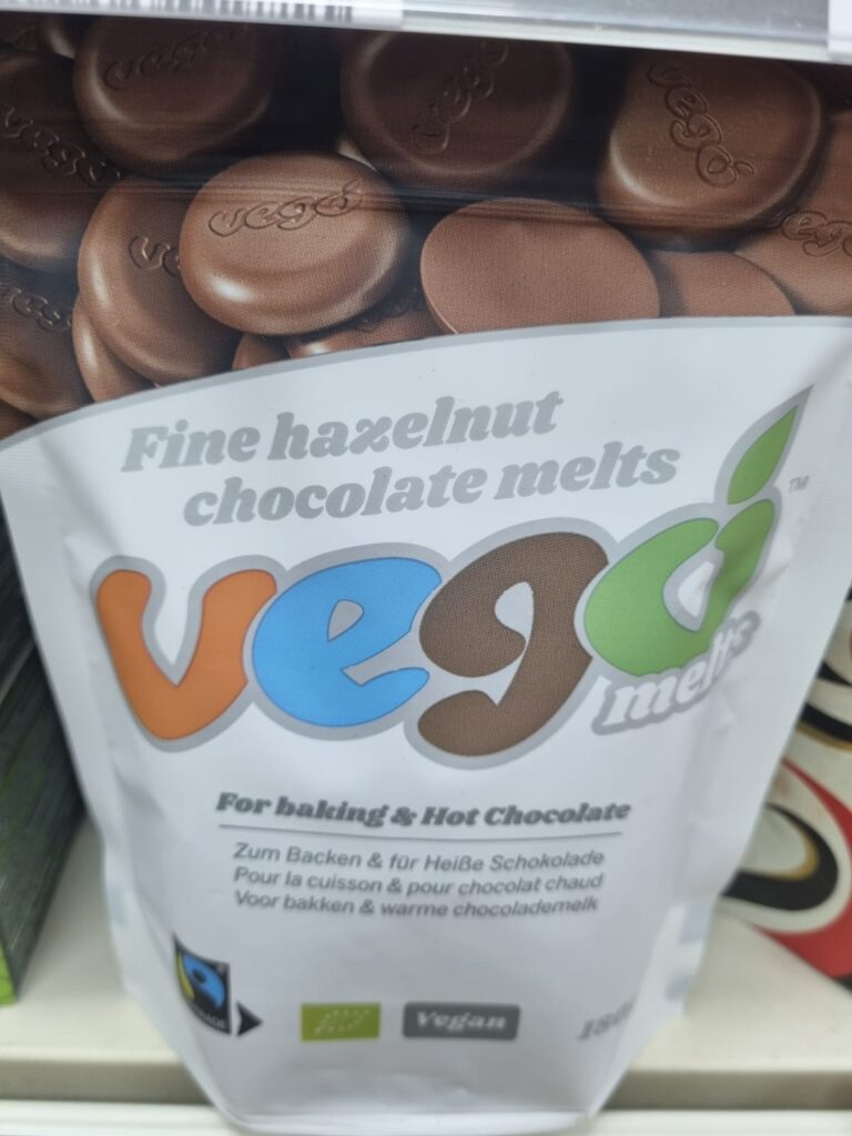 This image shows a bag of "Vego Melts," a product featuring fine hazelnut chocolate melts. The packaging is marked for use in baking and hot chocolate, and the product is labeled as vegan. The front of the bag includes multilingual text, specifying "For baking & Hot Chocolate" in English, along with translations in German, French, and Dutch. The package also displays fair trade and organic certifications. The image prominently features the "Vego" brand logo, with chocolate melts visible in the background.