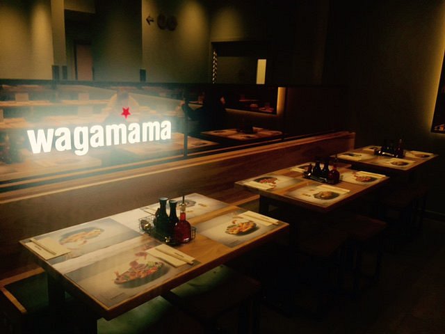 A dimly lit interior shot of Wagamama, showing neatly arranged tables with placemats and condiments. The restaurant's name is illuminated on a glass partition that separates the seating area.