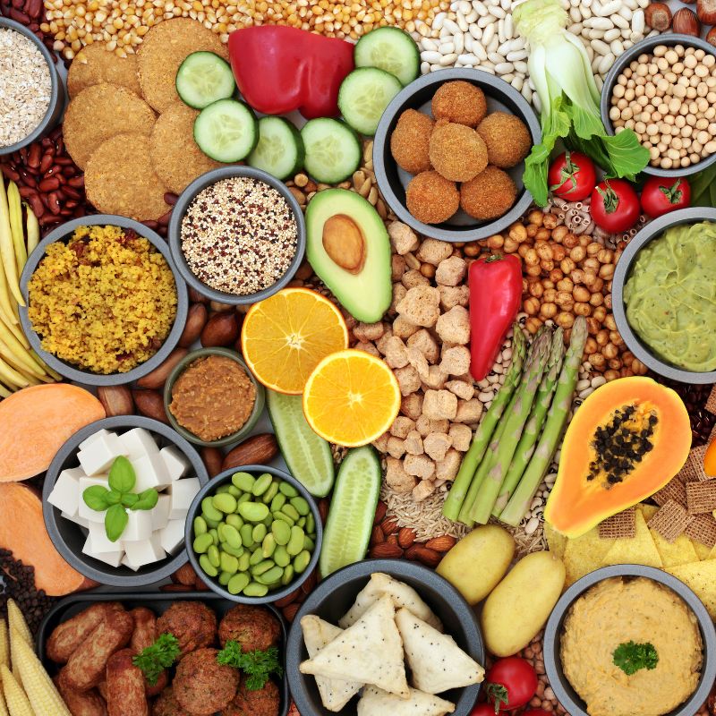 A vibrant assortment of plant-based foods, such as quinoa, lentils, chickpeas, tofu, fresh produce, and grains, arranged in small bowls and scattered across a light background.