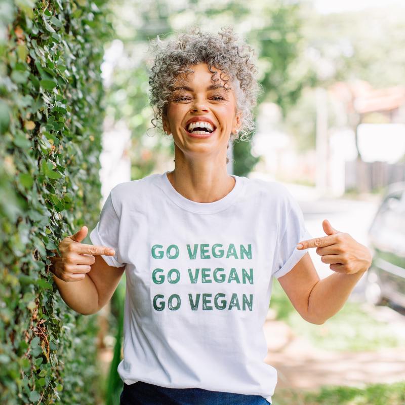 A smiling woman points at her white t-shirt, which has the phrase "Go Vegan" repeated three times in green letters. She stands outdoors near a leafy green wall, radiating enthusiasm.