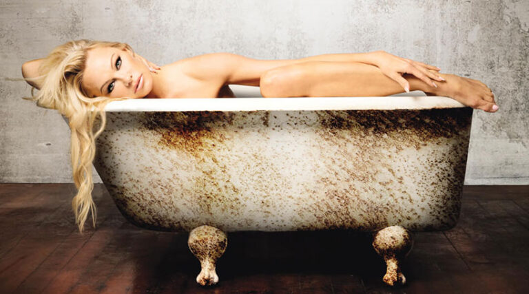 Pamela Anderson lying in an aged, rust-stained clawfoot bathtub, posed artistically against a textured wall, with long blonde hair cascading over the tub's edge.