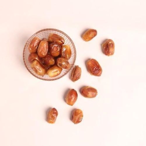  A small glass bowl filled with dates, with several dates scattered around it on a pink background