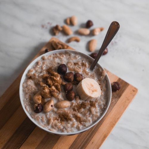 A bowl of oatmeal topped with a banana slice, walnuts, hazelnuts, and cinnamon, placed on a wooden board. A spoon is inside the bowl, and some nuts are scattered on the marble-like surface