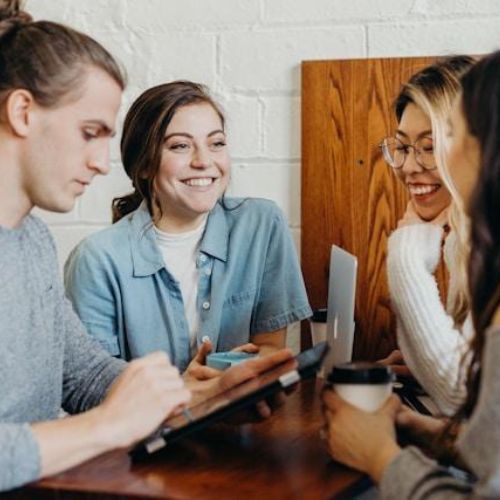 Four young adults sitting at a wooden table in a collaborative and friendly atmosphere. They are engaged in conversation and using tablets and coffee cups