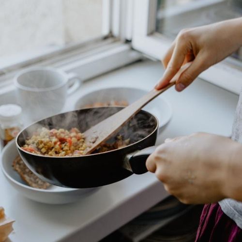 A person cooking on a stovetop, using a wooden spoon to stir a colorful vegetable stir-fry in a black frying pan