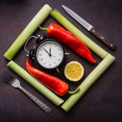 An arrangement of fresh vegetables and an alarm clock, symbolizing the importance of timing in nutrition or meal planning. The setup includes celery sticks, red bell peppers, a lemon slice, a fork, and a knife against a dark background