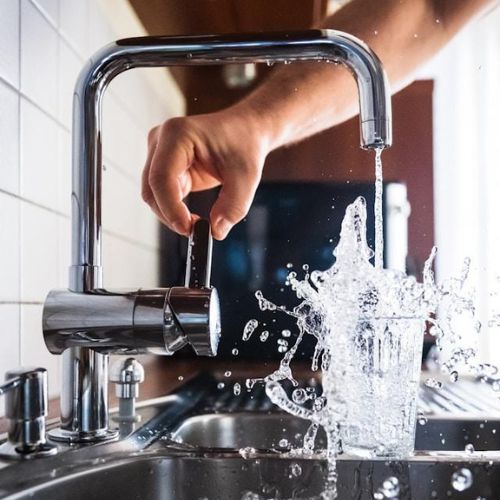 A person turning on a kitchen faucet, causing water to splash dramatically into a glass beneath it