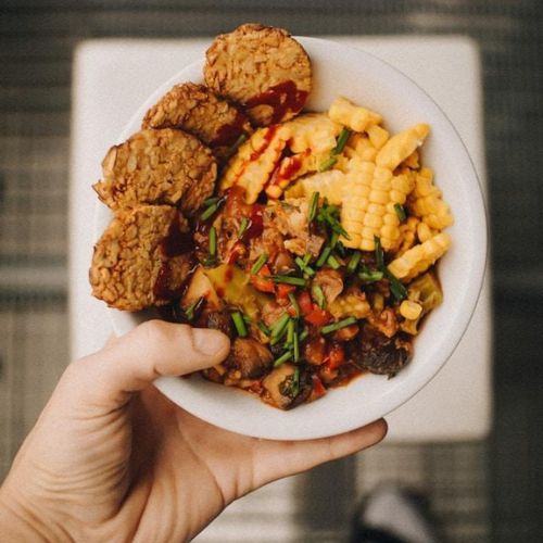 An overhead view of a person holding a bowl filled with a colorful and diverse meal. The bowl contains fried tempeh slices, corn, fries, and a mix of chopped vegetables garnished with chives