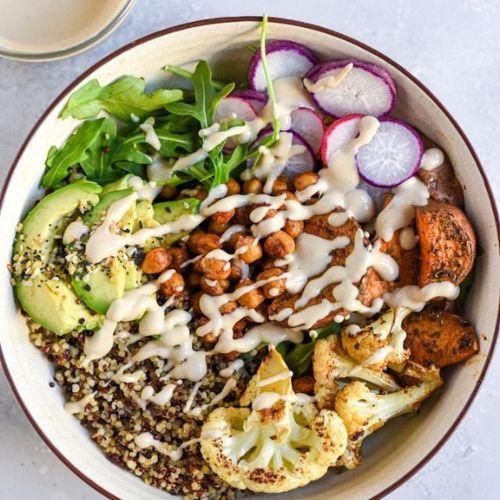 A bowl filled with a colorful arrangement of healthy ingredients, including quinoa, avocado slices, roasted chickpeas, arugula, radishes, roasted sweet potatoes, and cauliflower