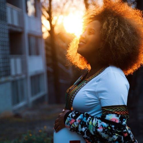 A pregnant woman standing outdoors in the evening, holding her belly and smiling, with the soft glow of a sunset in the background