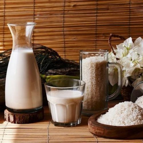 A rustic scene featuring a wooden table adorned with rice and rice milk. There is a glass pitcher and a smaller glass filled with rice milk, and a container overflowing with rice grains