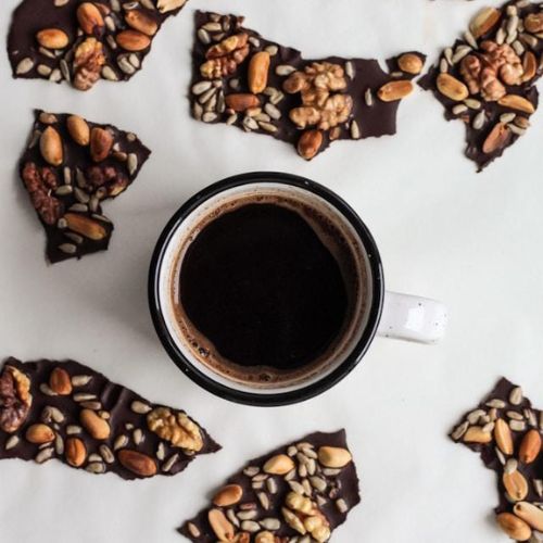 A cup of black coffee surrounded by pieces of dark chocolate bark topped with various nuts and seeds