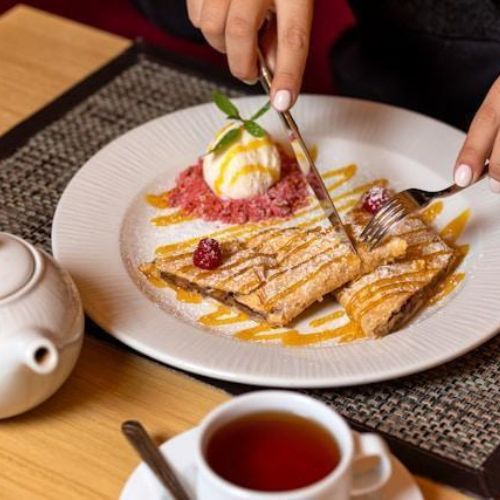 A person enjoying a dessert of crepes topped with ice cream, fresh raspberries, and a drizzle of caramel sauce. They are cutting into the crepe with a fork and knife