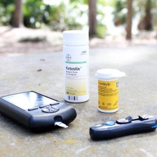 A selection of diabetes management tools laid out on a stone surface. The items include a glucose meter, a lancing device, and bottles of test strips (Ketostix and Freestyle)