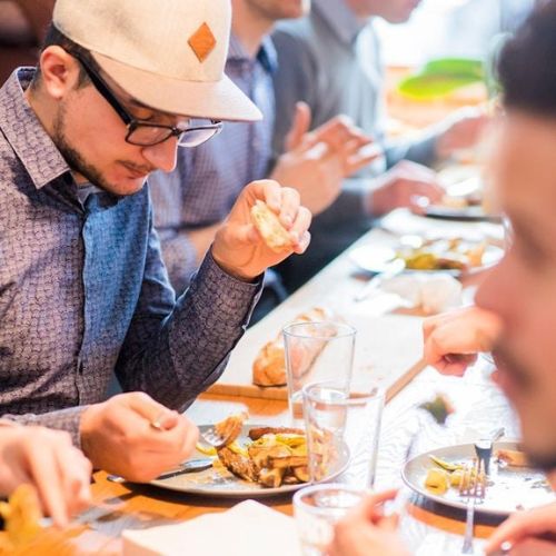 A man seated at a long table with other diners, enjoying a meal. He is focused on his dish, taking a bite of bread