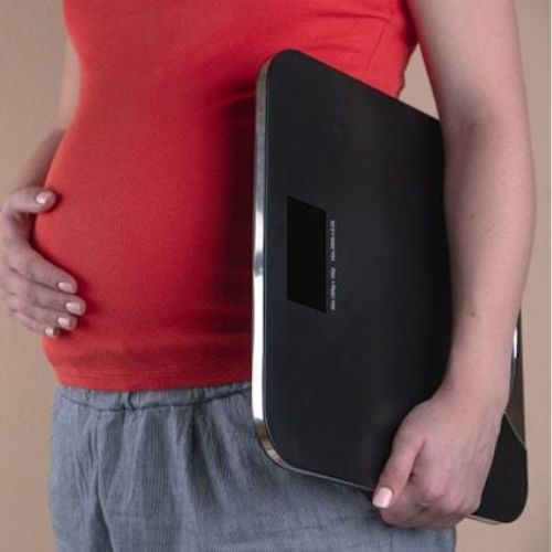 A pregnant woman wearing a red top and blue pants, holding a black digital scale against her side