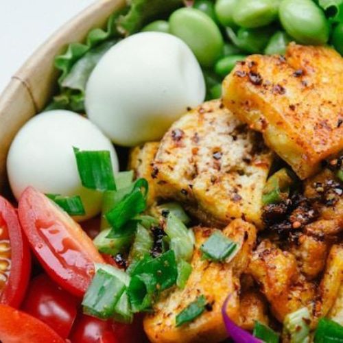 A close-up of a vibrant and healthy salad bowl. The bowl includes grilled tempeh cubes, cherry tomatoes, green onions, corn, edamame, and boiled eggs