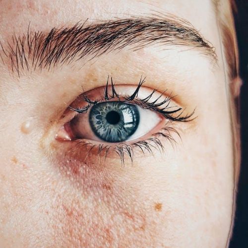 A close-up of a person's blue eye, highlighting its intricate details, including the pupil, iris, and eyelashes. The image also shows the texture of the surrounding skin, including freckles, pores, and a small beauty mark