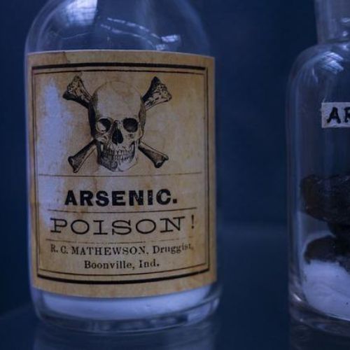 Two vintage glass bottles labeled "Arsenic." One bottle has a yellowed label with a skull and crossbones, marked "Poison." The other bottle has black crystals and is simply labeled "Arsenic."