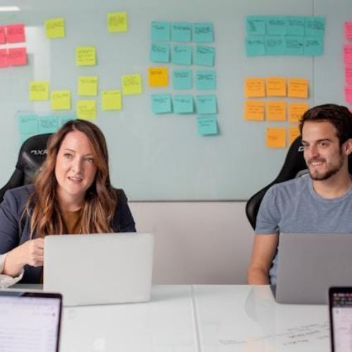 A group of people in a meeting room, with a whiteboard full of colorful sticky notes in the background. A woman in a blazer sits at the center with a laptop, actively engaging in a discussion, while a man in a casual shirt next to her also participates